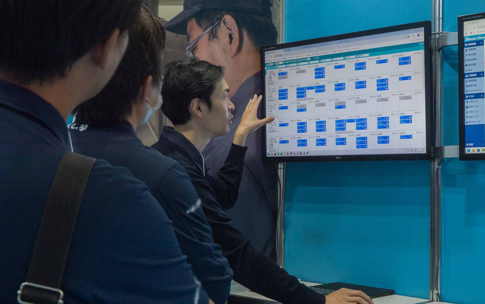 展示会ブース内で、スタッフが来場者に対し、モニターに映し出された進捗管理システム「アシストボード」のスケジュール画面を指差しながら説明している様子。