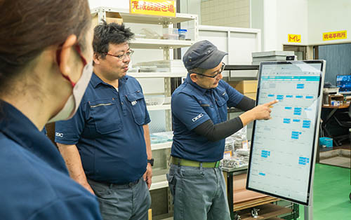 工場内に設置された大型モニターで生産計画のガントチャートを見て話し合う現場スタッフ